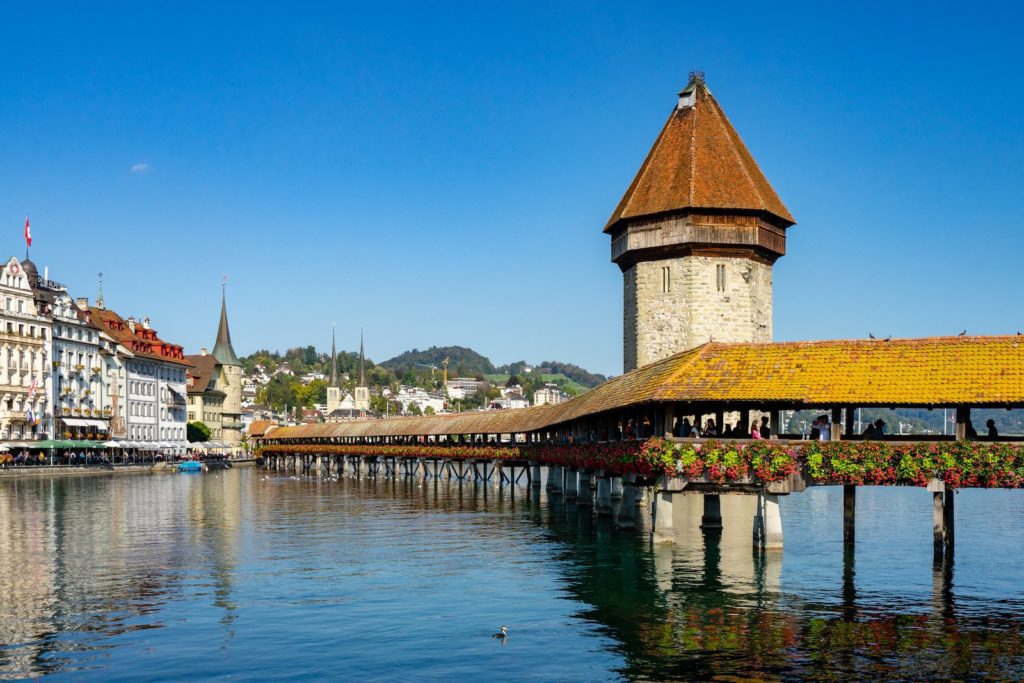 This image shows the Chapel Bridge and Water Tower in Lucerne, Switzerland, reflecting in clear water with colorful flowers and traditional architecture.
