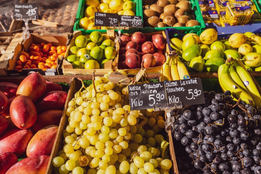 This image depicts a vibrant fruit market stall with various fruits, such as grapes, bananas, and mangoes, priced and labeled in euros per kilo.