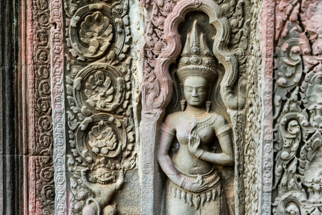 A detailed stone carving depicts an elegantly poised figure surrounded by intricate floral patterns on a temple wall, symbolizing historical or religious significance.