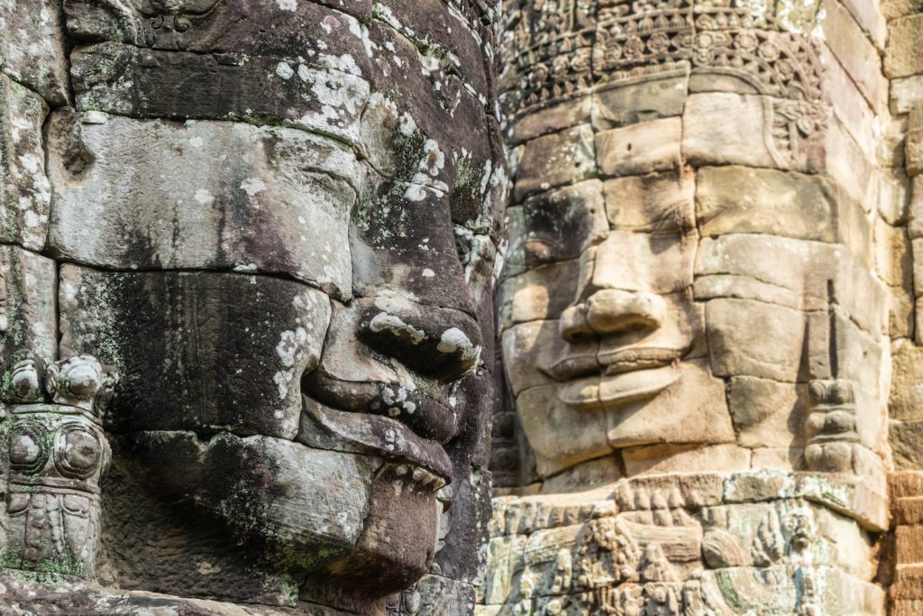 Two massive, serene stone faces, possibly representing Bodhisattvas, are carved into the facade of an ancient temple, weathered by time and overgrown with vegetation.