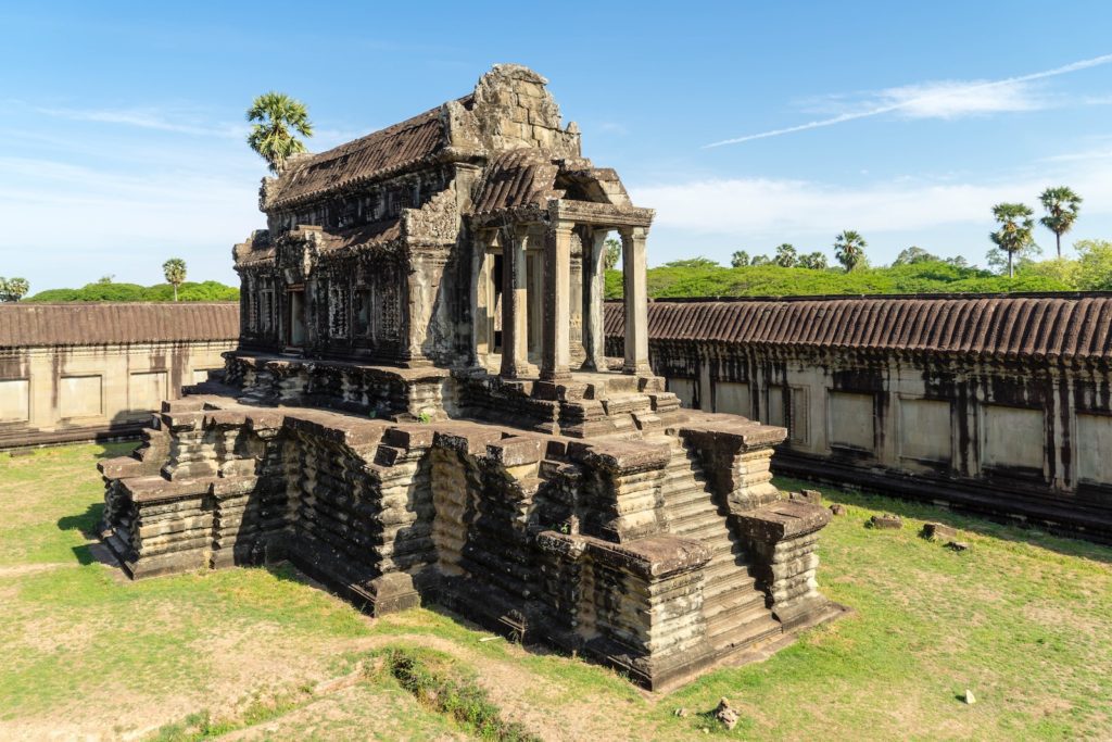 An ancient, weathered temple with intricate carvings stands amidst a grassy courtyard, surrounded by a long, colonnaded corridor under a clear blue sky.