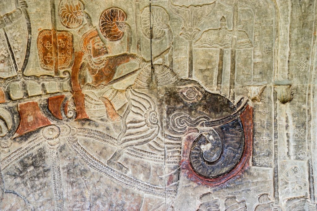 This image features an ancient, detailed bas-relief carving with traces of colored paint, depicting an elephant and two figures, possibly from a historical or mythological scene.