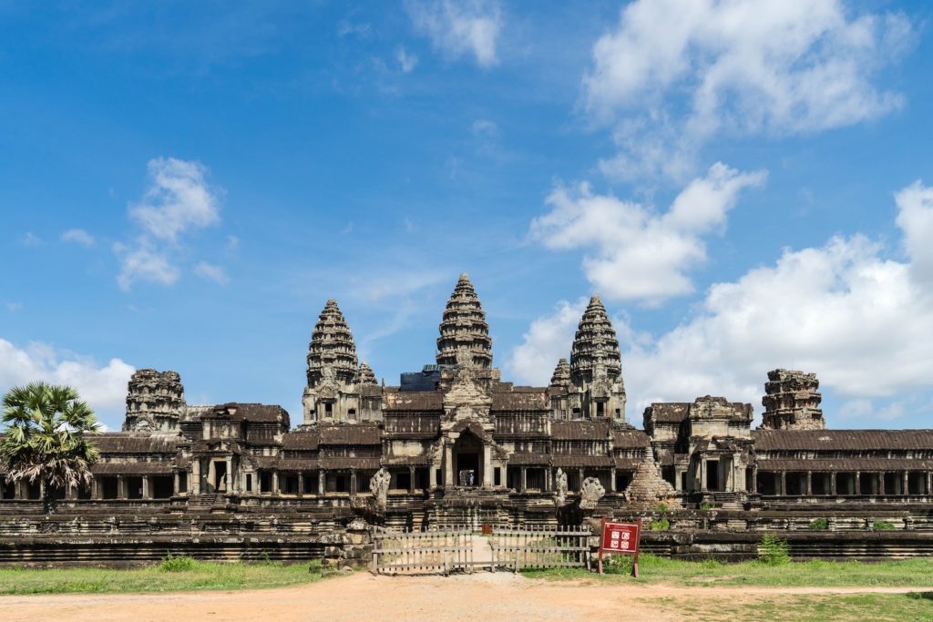 The image shows the ancient temple complex of Angkor Wat in Cambodia, featuring its iconic towers, intricate stone carvings, and a clear blue sky.