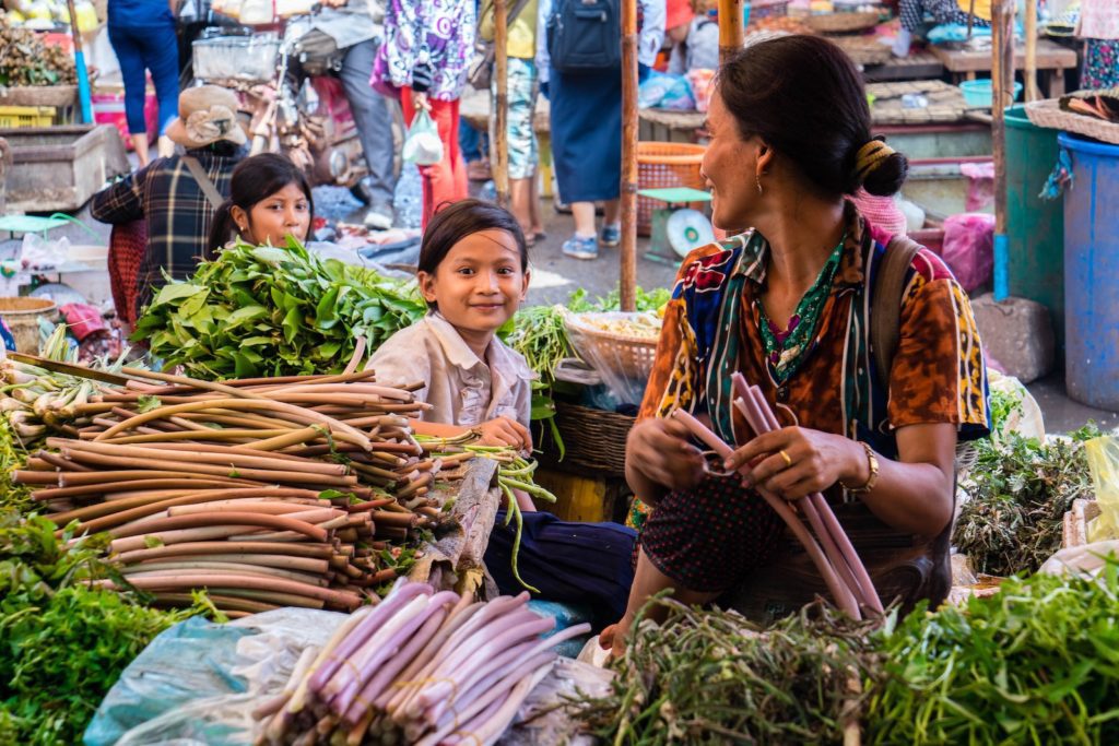 A bustling market scene where a person is selling fresh vegetables with a child beside them smiling and various shoppers walking in the background.