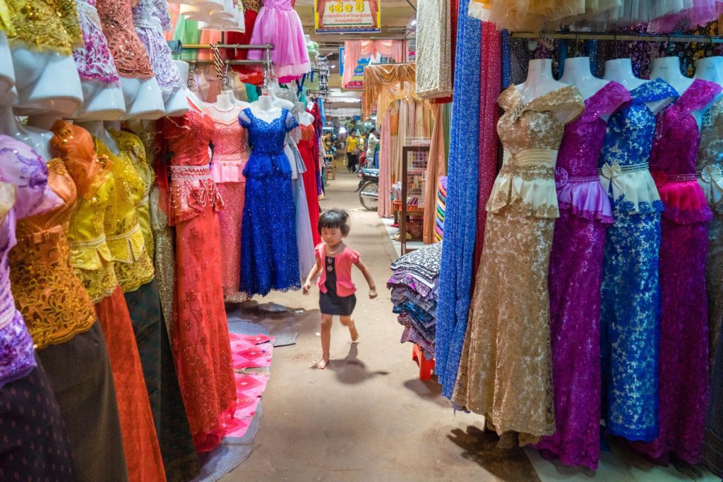 A young child walks through a colorful marketplace aisle lined with elaborate dresses on hangers, creating a vibrant, textured scene of fabrics and patterns.