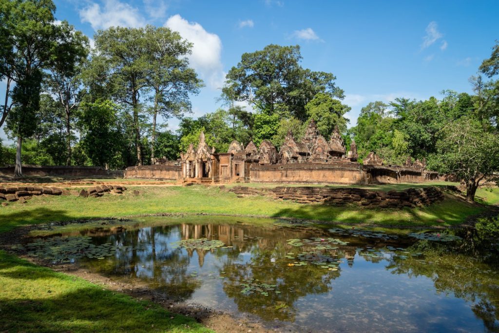 Ancient temple ruins stand amidst green trees, reflected in a tranquil water pond under a blue sky with fluffy clouds. A serene historic landscape.