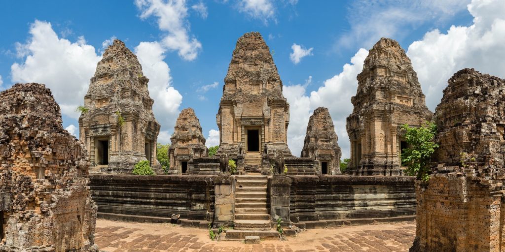 The image displays an ancient temple complex with weathered stone towers and ruins under a partly cloudy sky, showcasing historical architecture and cultural heritage.