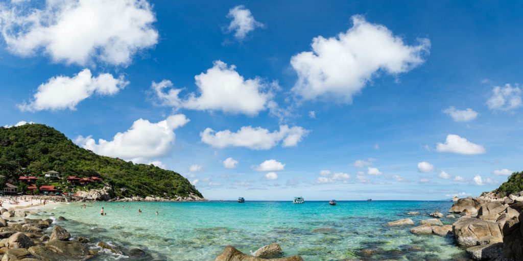 This image captures a panoramic view of a tropical beach with clear blue waters, people swimming, scattered clouds in the sky, and rocky foreground.