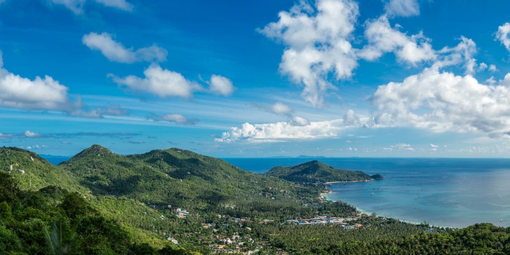 A panoramic view featuring lush green hills descending toward a coastline with buildings, under a vast, partly cloudy sky above a serene blue sea.