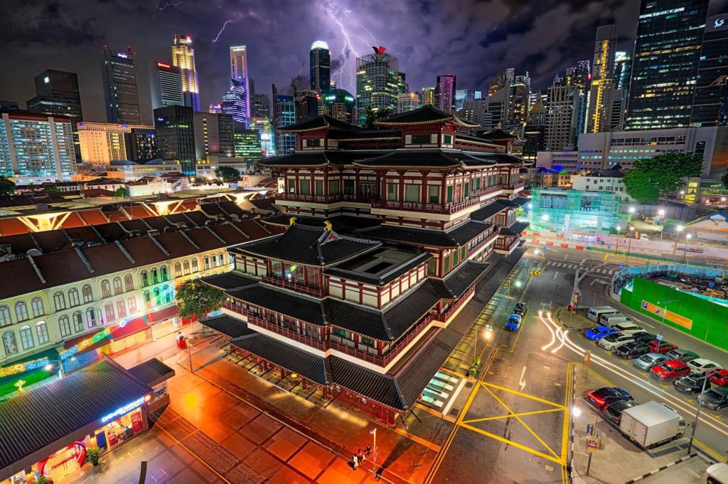 A vibrant nighttime cityscape showing a traditional multi-tiered building contrasting with modern high-rise skyscrapers in the background, illuminated streets, and a busy parking lot.