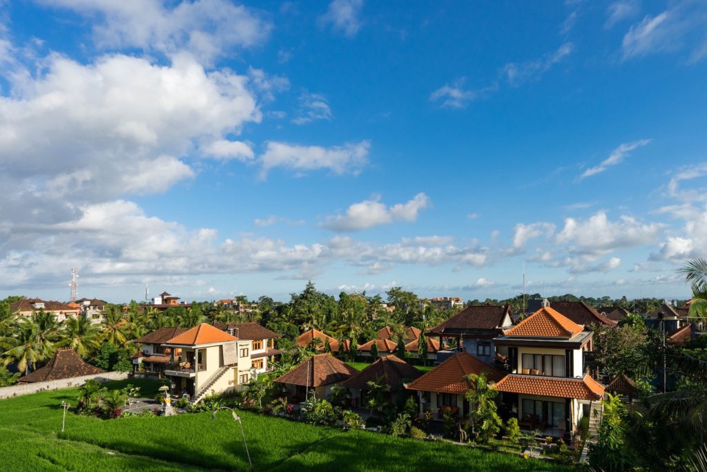 Lush green rice terraces in the foreground with traditional Balinese-style houses amidst abundant tropical vegetation under a blue sky with clouds.