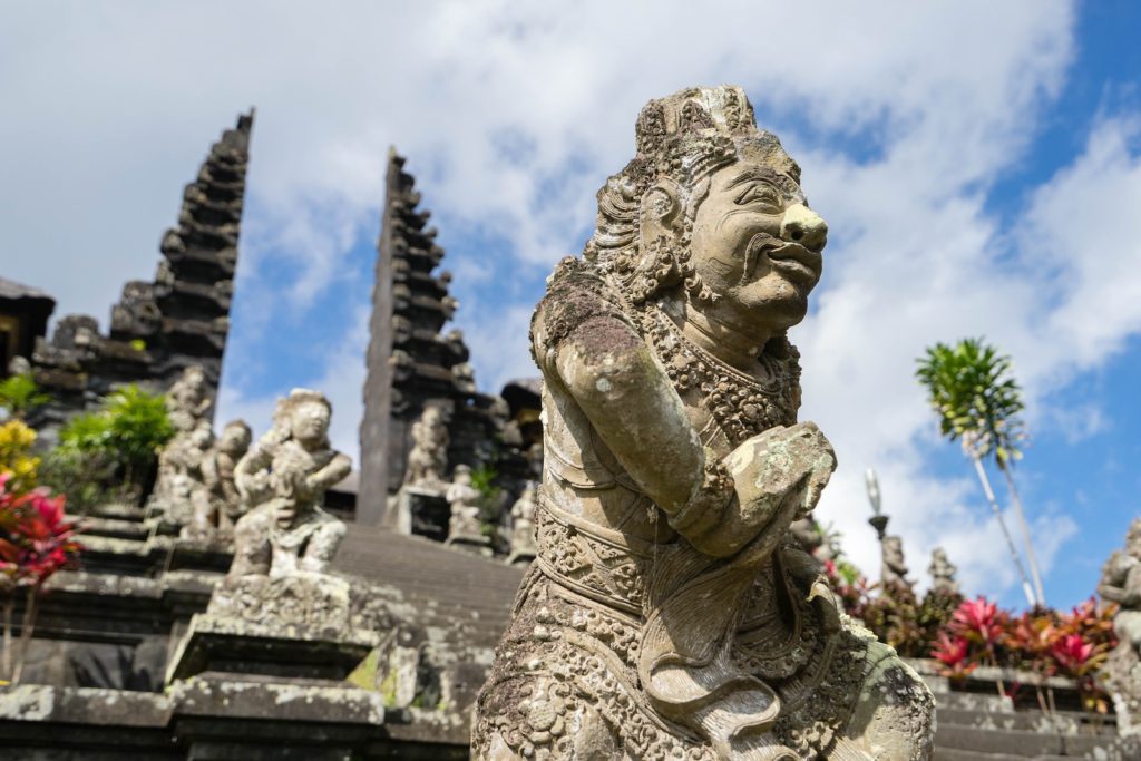 A stone sculpture with intricate carvings depicting a mythological figure stands in the foreground. Behind, tall, tiered temple towers rise into a blue sky with clouds.