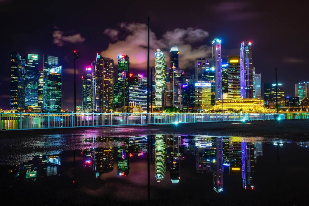 A nighttime cityscape with illuminated skyscrapers reflected in water. A vibrant display of neon lights creates a futuristic and dynamic urban atmosphere.