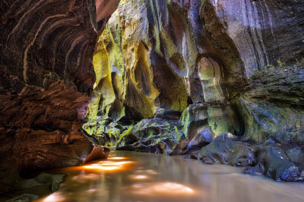 This image captures a serene slot canyon with flowing water reflecting light, surrounded by smooth, multicolored rock walls showing varied geological layers and textures.