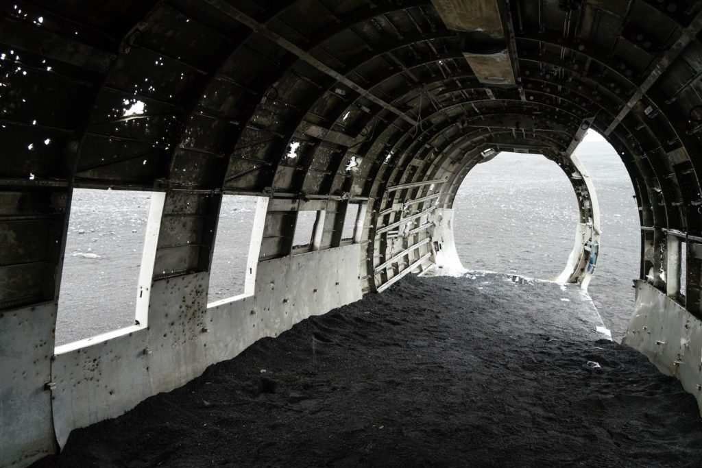 This image depicts an abandoned, weathered airplane fuselage with missing panels, partially filled with dark soil or sand, situated against a backdrop of overcast skies.