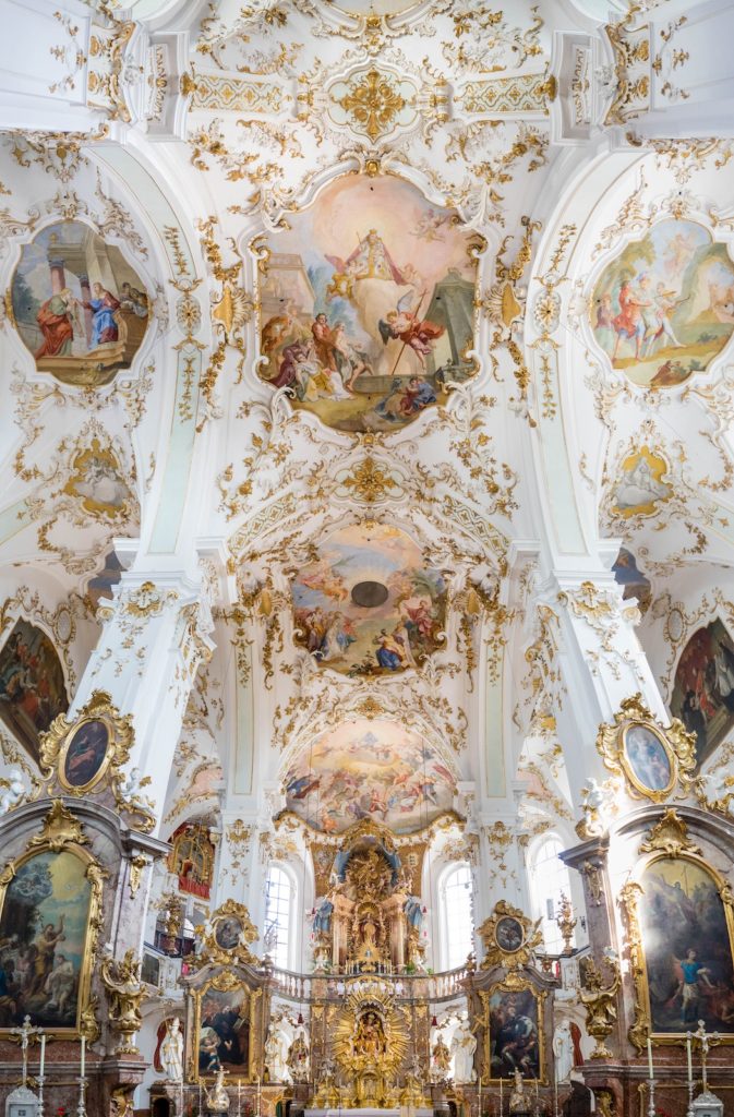 The image shows the ornate interior of a Baroque-style church with elaborate gold decorations, frescoes, and religious paintings on the walls and ceiling.