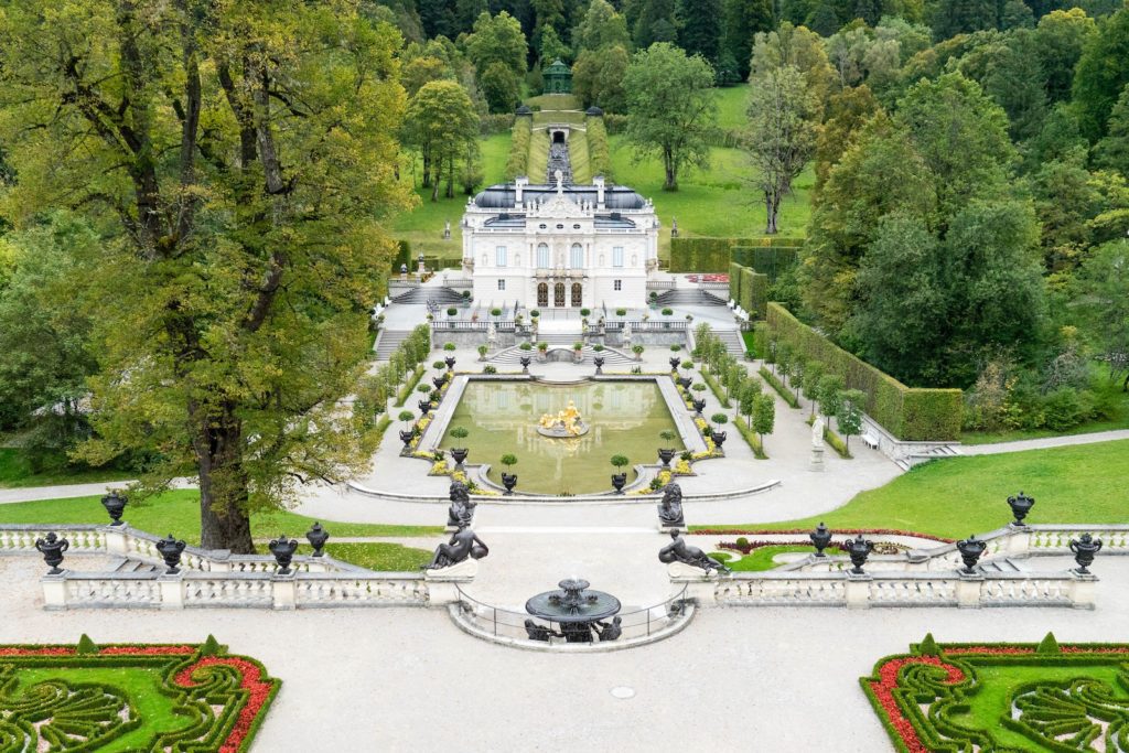 This image shows a grand estate surrounded by manicured gardens, symmetrical pathways, decorative statues, and hedges, with dense trees in the background.