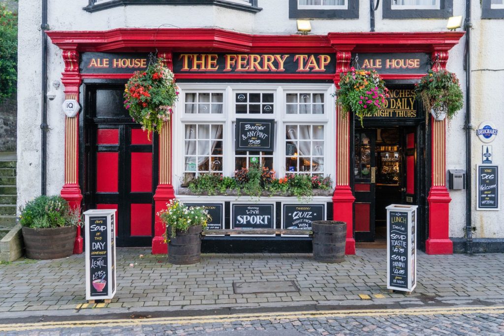 A traditional British pub, "The Ferry Tap," is adorned with red and black paint, flowering plants, and chalkboard signs promoting food and sports viewing.