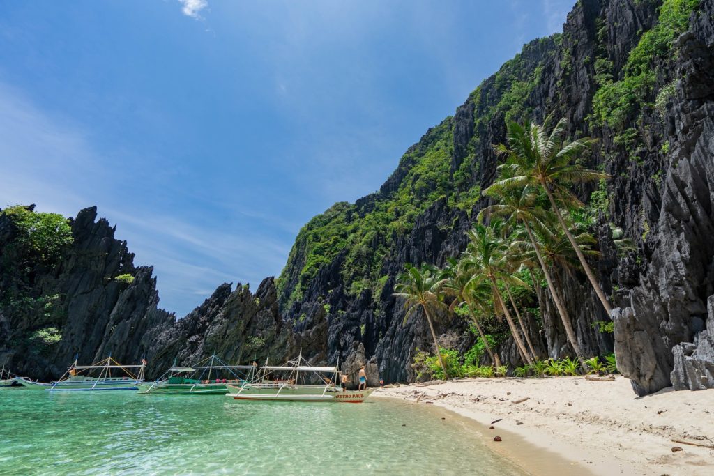 A tropical beach with clear water, traditional boats, a person, palm trees, and imposing cliffs under a blue sky with clouds. A serene vacation spot.
