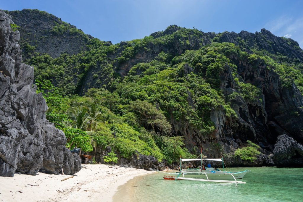 A tropical beach with pristine sand is flanked by lush greenery and rugged cliffs. A traditional boat floats near the shore under a clear blue sky.