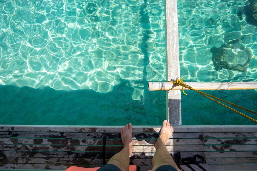 A person sits on a wooden dock over crystal-clear turquoise water, looking down at their bare feet, with a yellow rope tied nearby.
