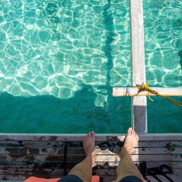 A person sits on a wooden dock over crystal-clear turquoise water, looking down at their bare feet, with a yellow rope tied nearby.