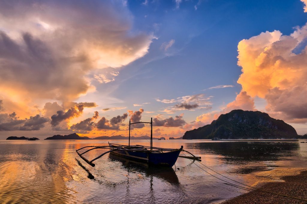 A traditional boat rests on a calm sea at sunset, with dramatic clouds in the sky and islands on the horizon creating a picturesque scene.