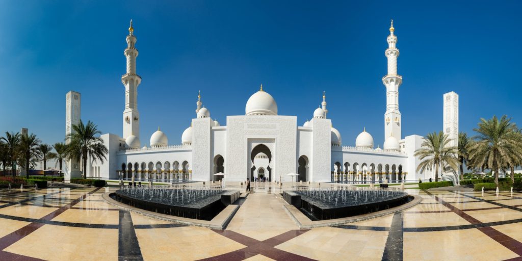 This image shows the Sheikh Zayed Grand Mosque with white domes, minarets, and a reflection pool under a clear blue sky. Palm trees flank the sides.