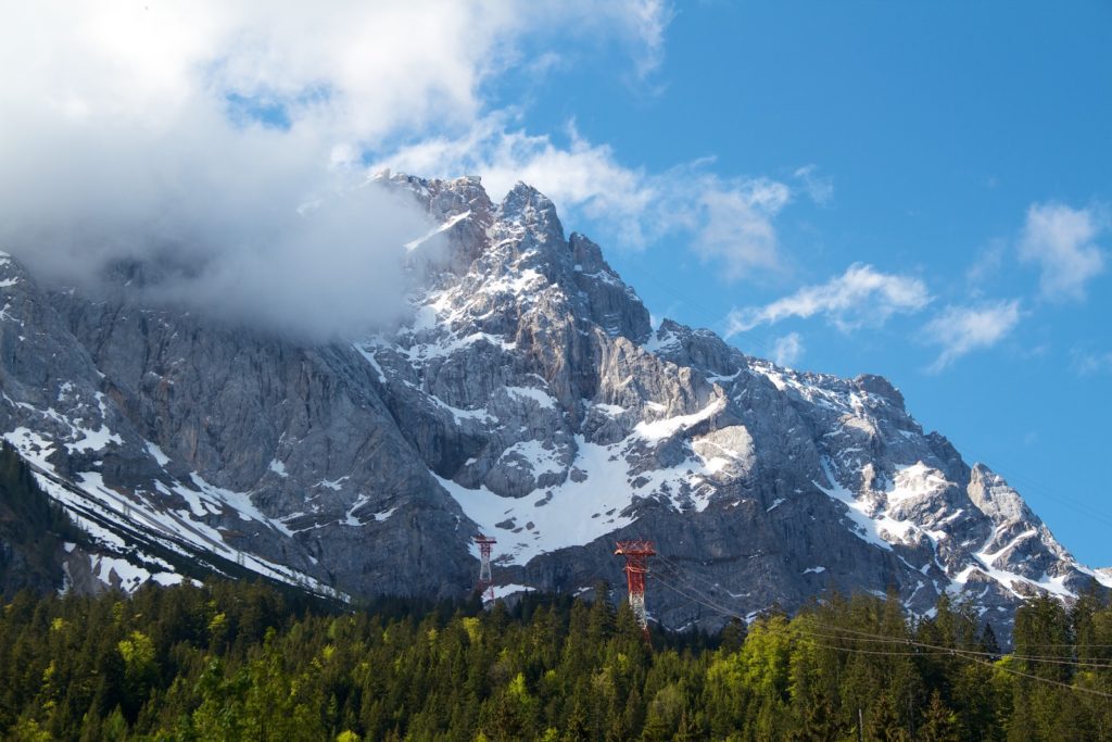 Majestic mountain peaks with snow patches tower above a lush green forest under a bright blue sky with wispy clouds and a cable car tower visible.