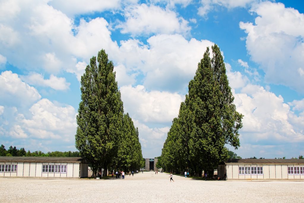 A symmetrical view between two large trees, under a cloudy blue sky, overlooking a wide courtyard with long, low buildings and scattered visitors.