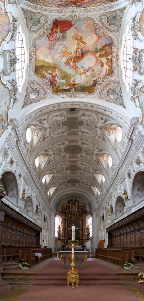 Baroque-style church interior with ornate fresco ceiling, intricate white stucco decorations, wood pews, and elaborate golden altar. A crucifix stands in the aisle.