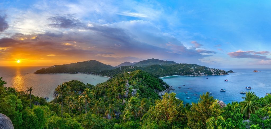 A panoramic view of a tropical coastline at sunset, with lush hills, boats on water, under a sky with clouds reflecting warm sunlight.