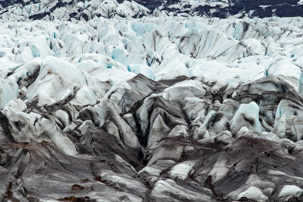 This image shows a rugged glacier with deep blue crevasses and dark sediment lines. The textured ice expanse contrasts with the underlying rock.