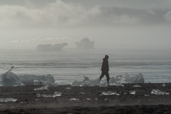 A person is walking on a misty beach scattered with transparent ice chunks; distant icebergs and foggy air convey a serene, cold atmosphere.