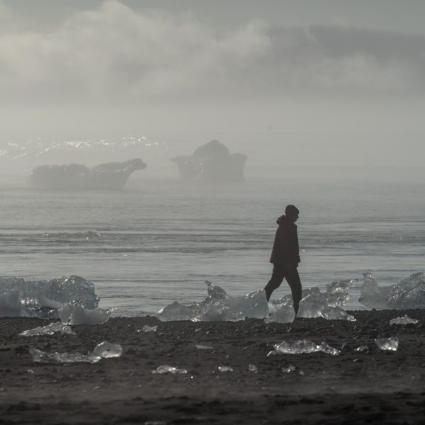 A person is walking on a misty beach scattered with transparent ice chunks; distant icebergs and foggy air convey a serene, cold atmosphere.