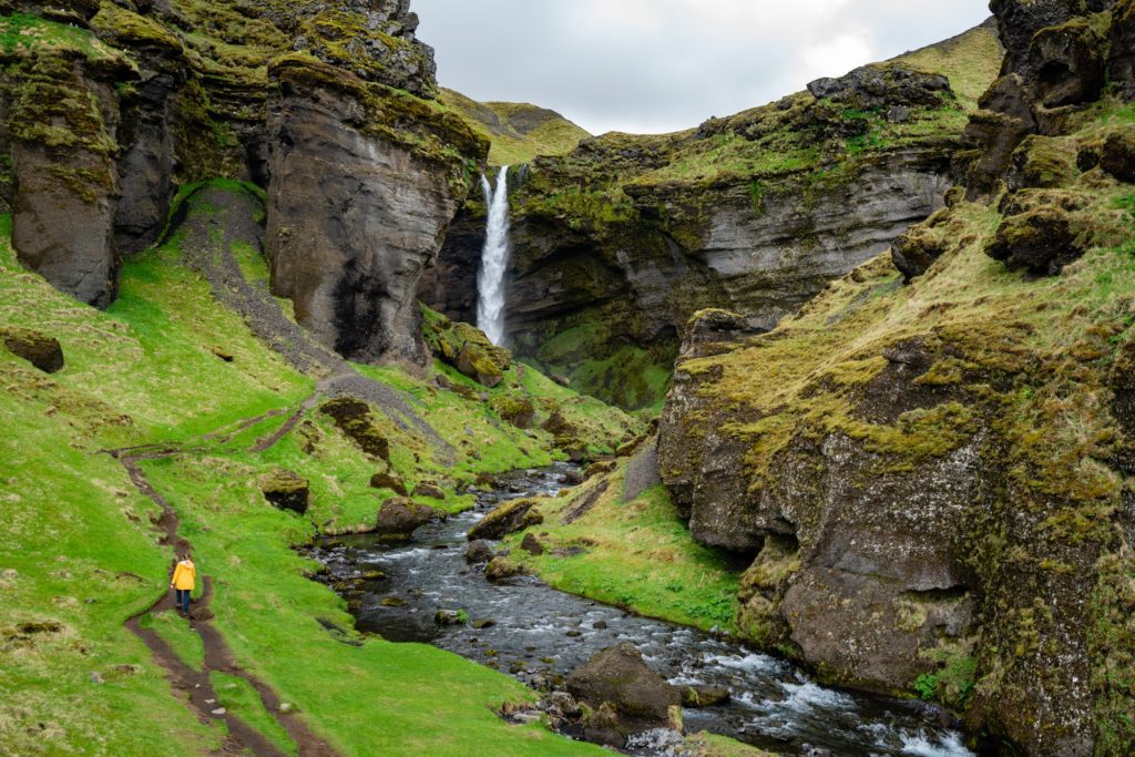 A person in a yellow coat walks on a path along a stream flowing from a waterfall within a lush, moss-covered canyon under a cloudy sky.