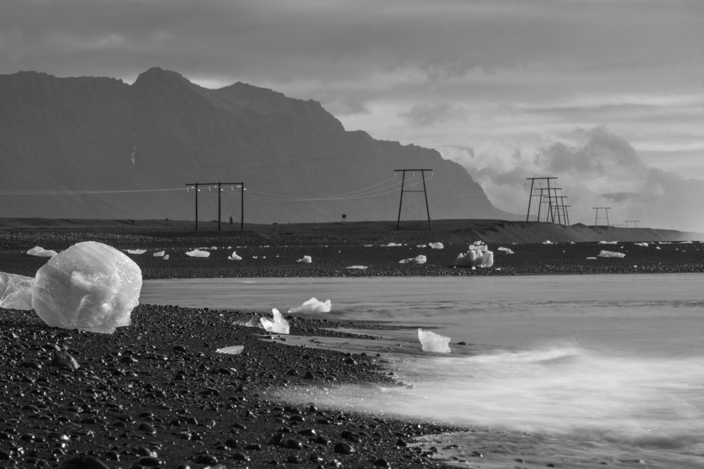 This black and white image shows a pebble beach with chunks of ice, gentle waves, electricity pylons in the distance, and mountainous terrain in the background.