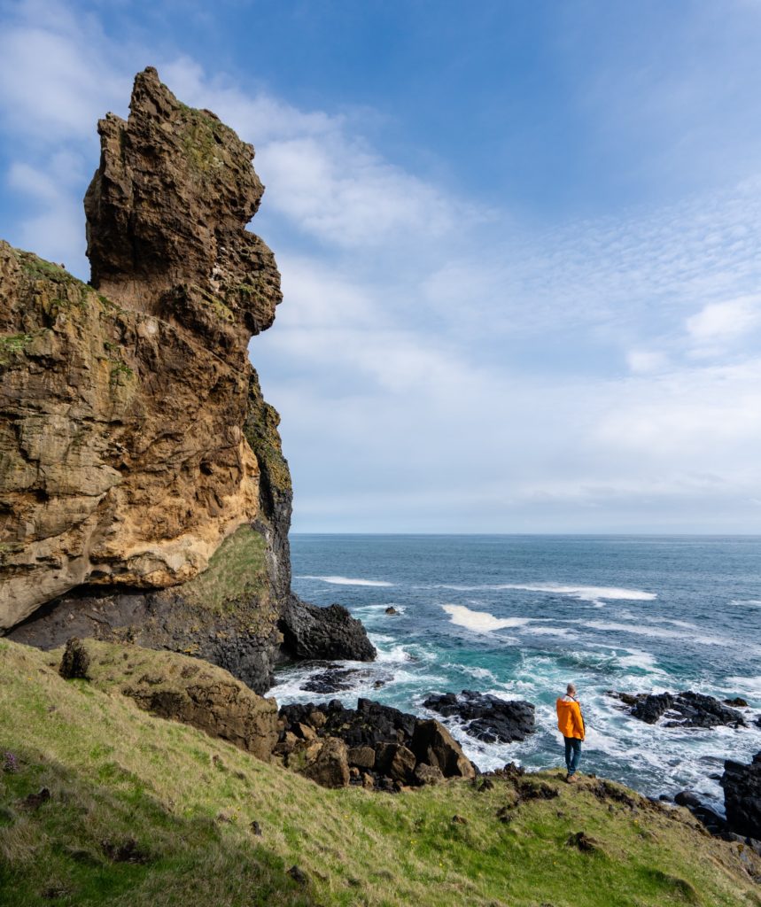 A person in an orange jacket stands on a grassy coastline, observing a vast ocean beside a towering rocky cliff under a blue sky with clouds.