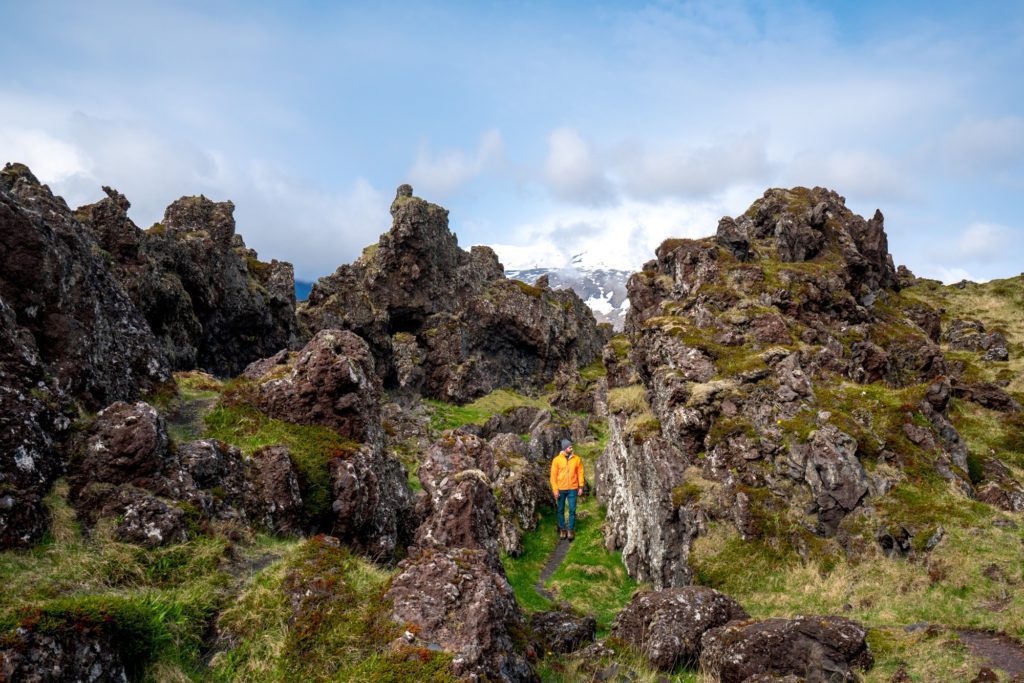 A person in an orange jacket stands on a trail amidst rugged, moss-covered rocks under a blue sky with white clouds and distant snowy mountains.