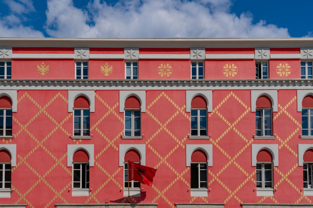 The image shows an ornate building façade with symmetrical windows, adorned with red and gold diamond patterns, and a flag fluttering against a blue sky.