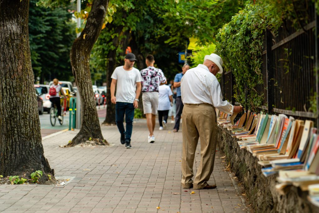 A person in a white hat browses books lined along an outdoor wall, while others walk by on a shaded pedestrian path flanked by trees.