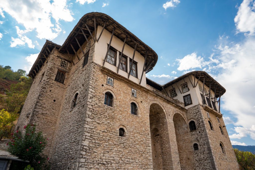 A traditional stone building with a prominent wooden upper structure stands against a blue sky with wispy clouds, nestled on a hillside with greenery.