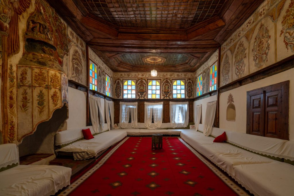 This is a traditional room featuring ornate wall decorations, wooden ceiling, colorful stained-glass windows, red carpet, low beds with white linens, and decorative pillows.