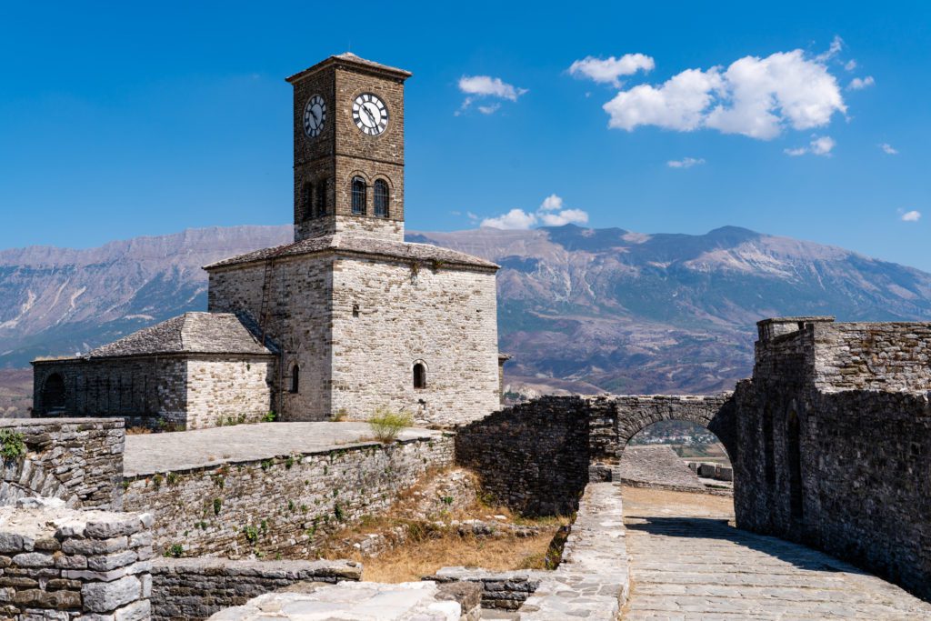 An old stone clock tower stands prominently within a historic fortress under a clear blue sky, with mountains in the background and wispy clouds above.