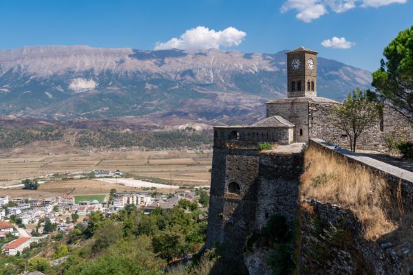 An ancient stone fortress with a bell tower overlooks a valley with buildings below. In the distance, a majestic mountain range rises under a blue sky.