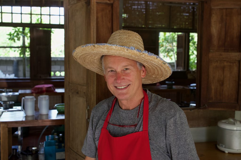 A smiling person wearing a straw hat and a red apron stands in a kitchen with wooden cabinetry and various appliances, exuding a pleasant demeanor.