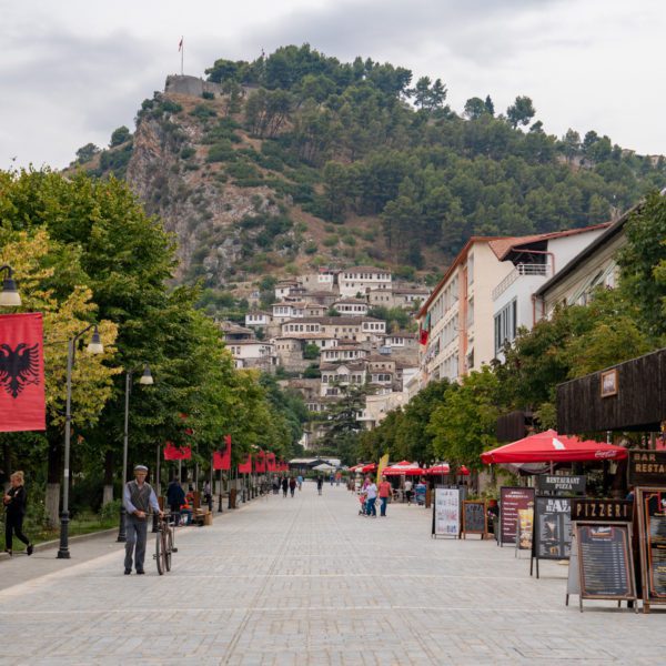 A pedestrian boulevard lined with trees and restaurants, banners, people walking, and a hill with buildings and a flag at the top in the background.