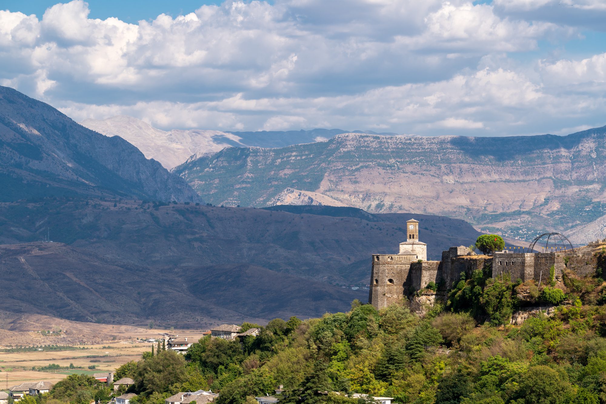 An ancient fortress with a prominent tower sits atop a hill, overlooking a valley with sprawling fields, backed by towering, cloud-dappled mountains.