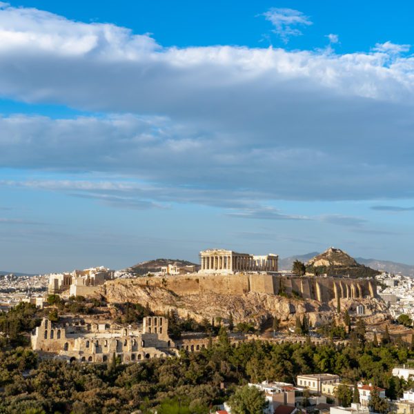 This image features the Acropolis of Athens, prominently displaying the Parthenon and surrounding ruins, with the sprawling cityscape beneath a blue sky with few clouds.