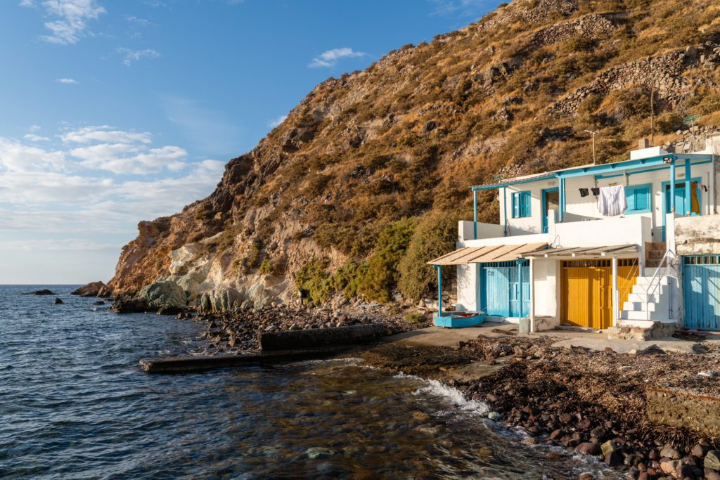 A quaint blue and white house sits on a rocky shoreline beside a clear sea, with clothing hanging outside and a steep hill looming behind.